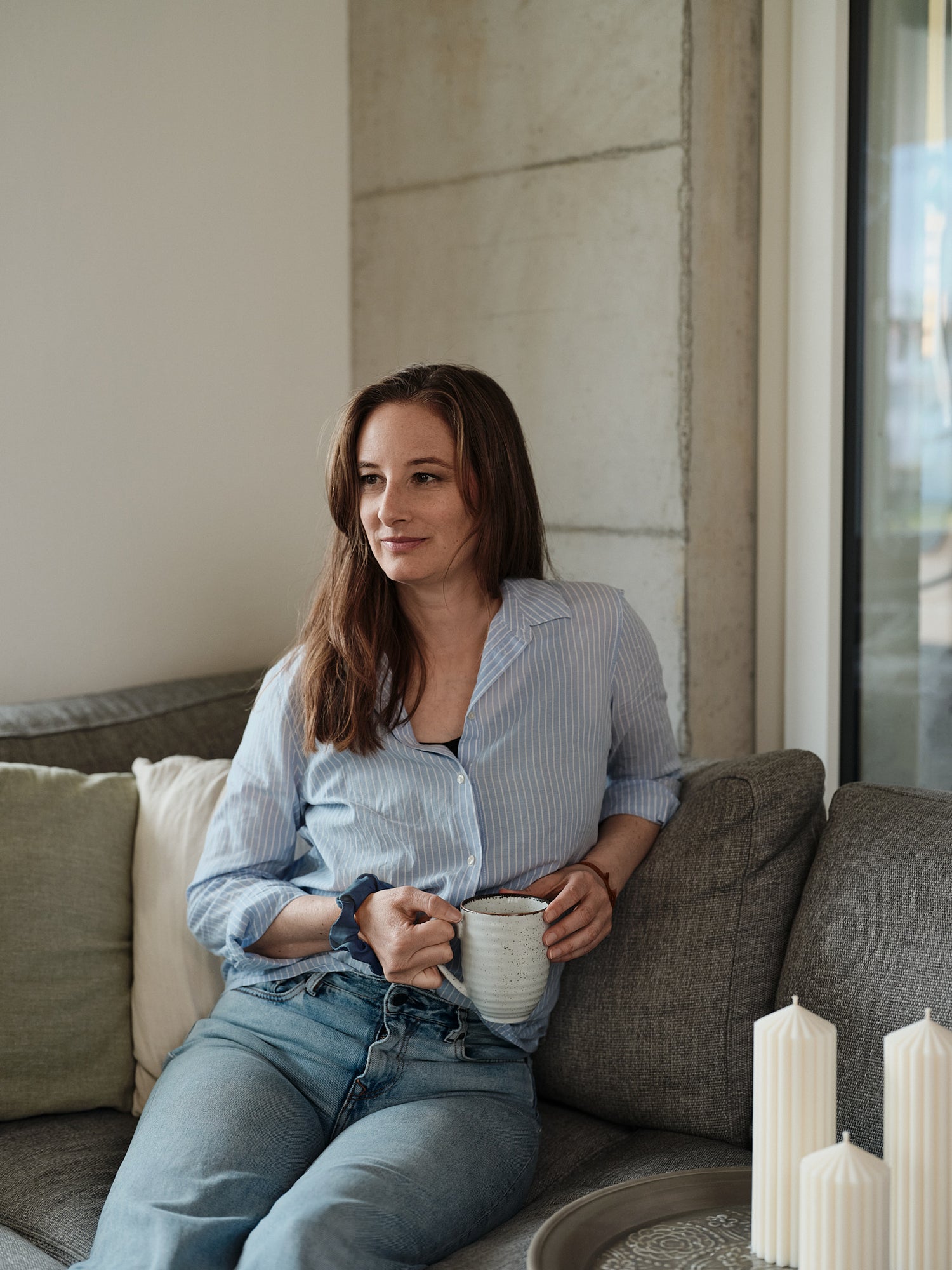 Jessica Niedermann, die Gründerin und Inhaberin von Olfair sitzend auf einem grauen Sofa mit Tee in der Hand und Zylinderkerzen als Dekoration