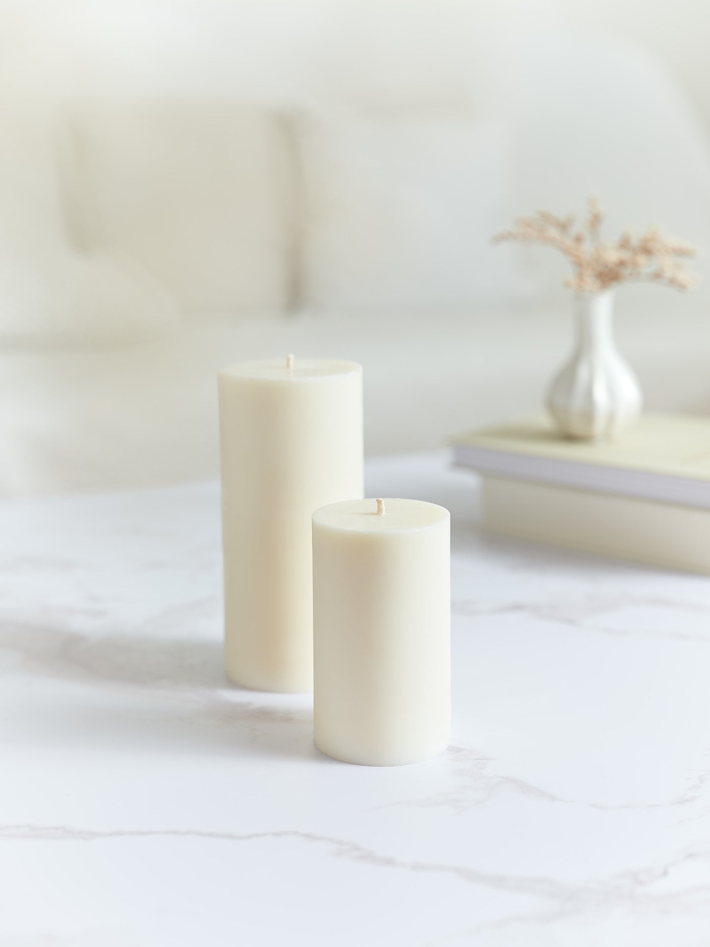 Eine grosse und eine kleine glatte Zylinderkerze in cremeweiss. Beide stehen auf einem Marmorcouchtisch und im Hintergrund hat es Bücher und eine Vase mit Trockenblumen.